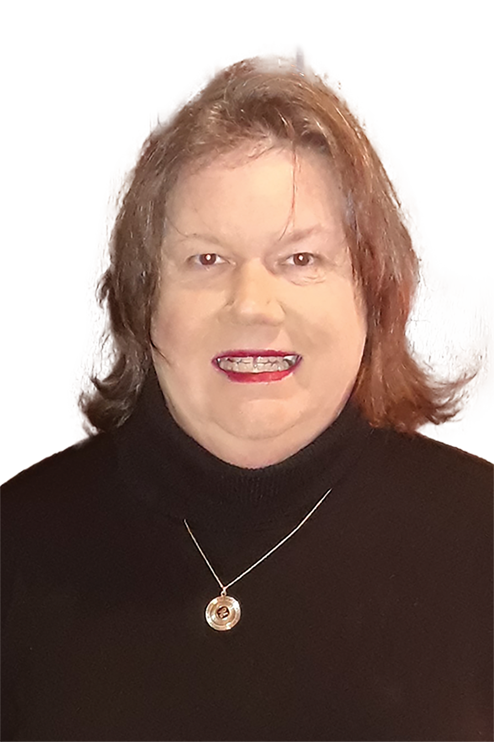 A trans middle-aged female with light brown hair, wearing a black long-sleeved turtleneck dress standing in front of a wooden door with a lamp in the background.