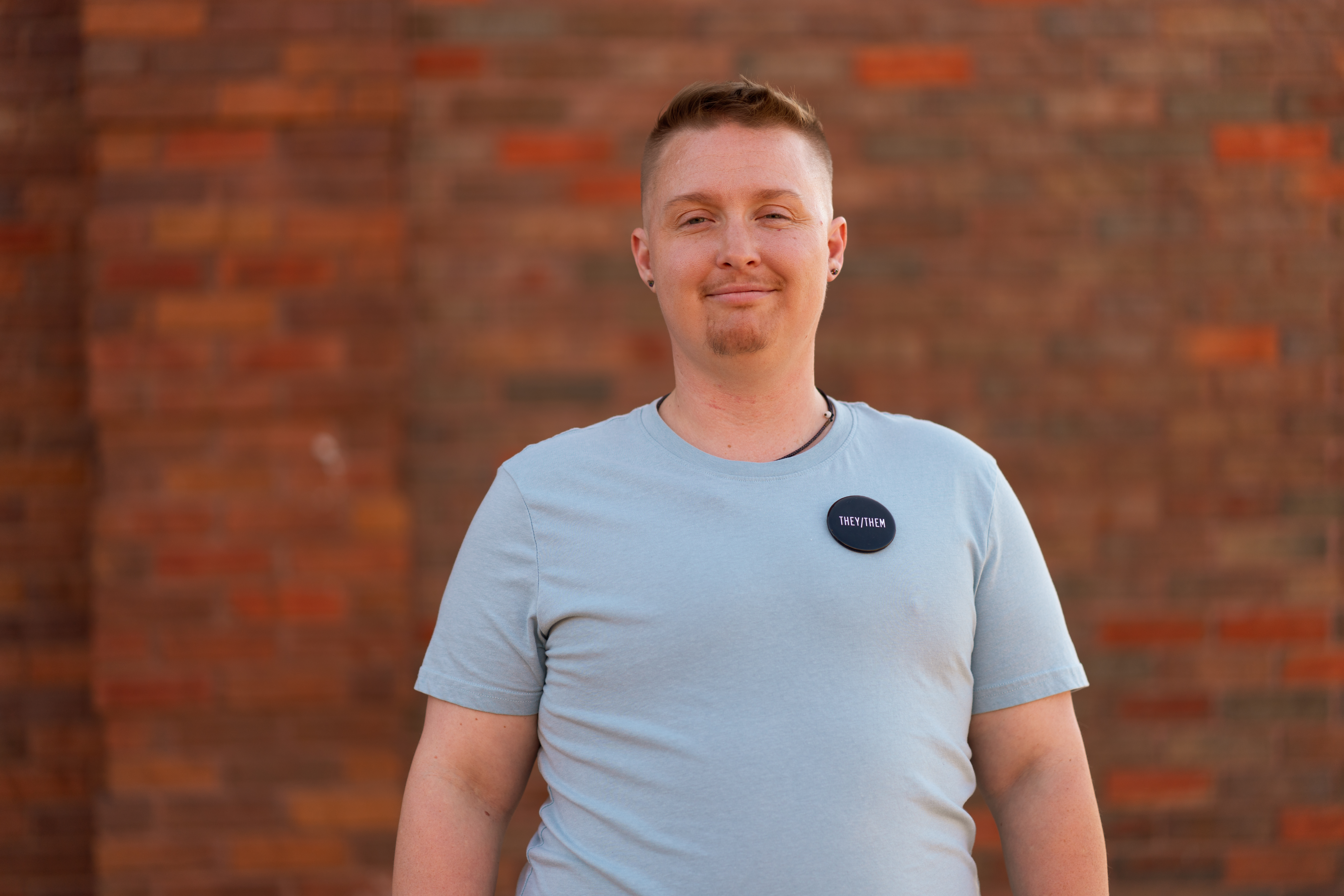 Nonbinary Arkansan wearing They/Them pin on blue t-shirt standing in front of red brick background
