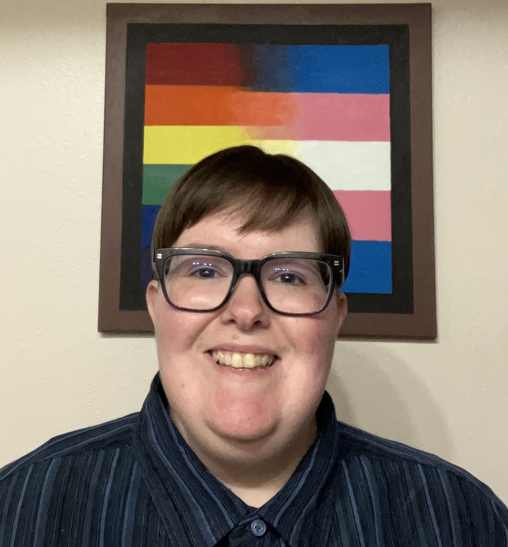 A smiling nonbinary Arkansan with short brown hear wearing a black striped button-down shirt and black glasses in front of a painting of a pride progress flag.