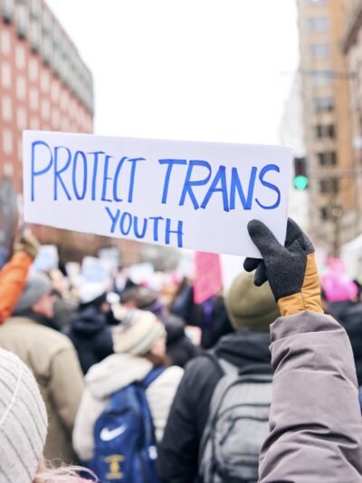 Protestor at a rally holding a handwritten sign in blue ink on white paper that reads: Protect Trans Youth