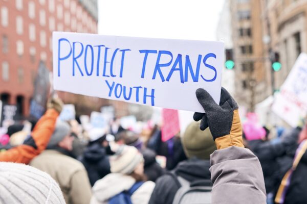 Protestor at a rally holding a handwritten sign in blue ink on white paper that reads: Protect Trans Youth
