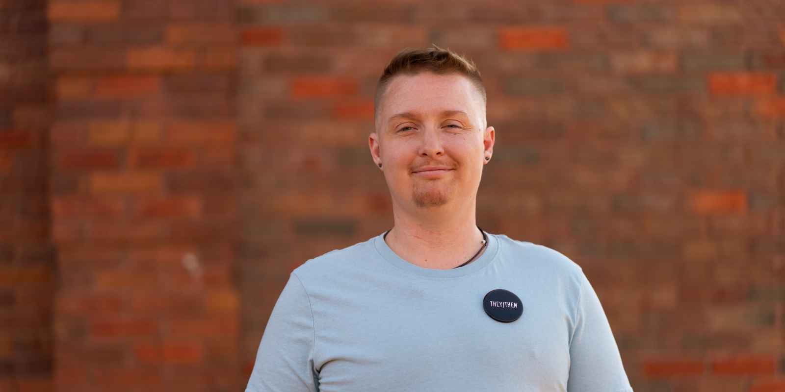 Nonbinary Arkansan wearing They/Them pin on blue t-shirt standing in front of red brick background