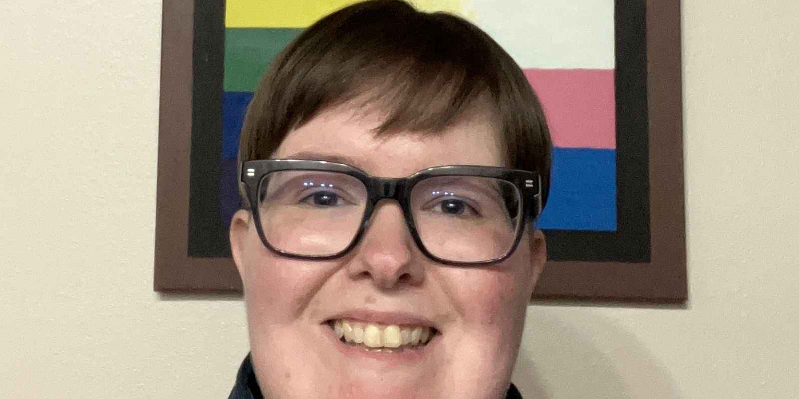 A smiling nonbinary Arkansan with short brown hear wearing a black striped button-down shirt and black glasses in front of a painting of a pride progress flag. 