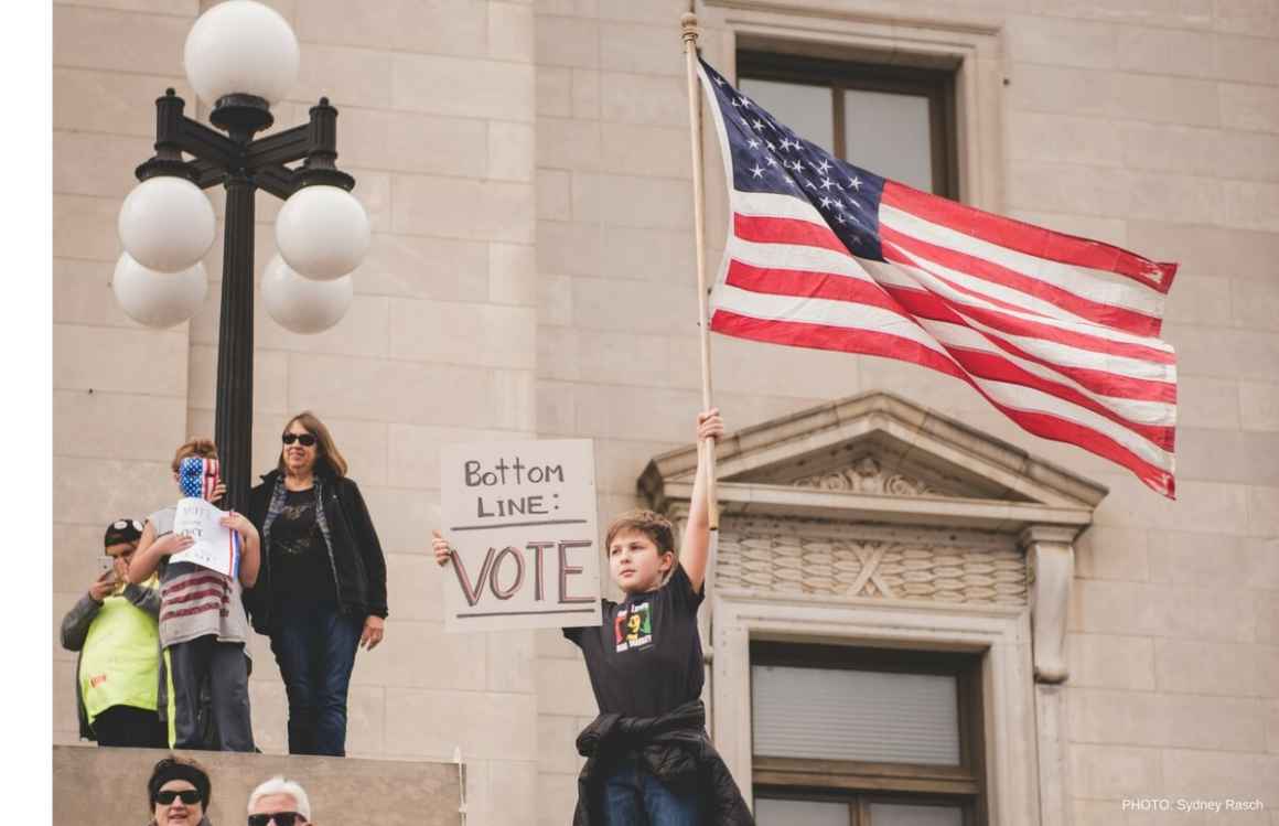 Boy holding an American flag and a sign that reads "Bottom Line: VOTE"