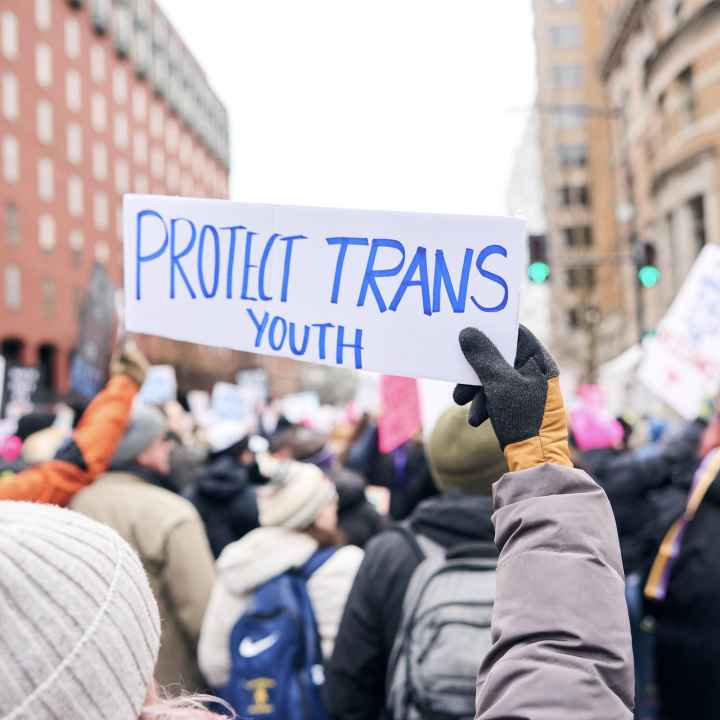 Protestor at a rally holding a handwritten sign in blue ink on white paper that reads: Protect Trans Youth